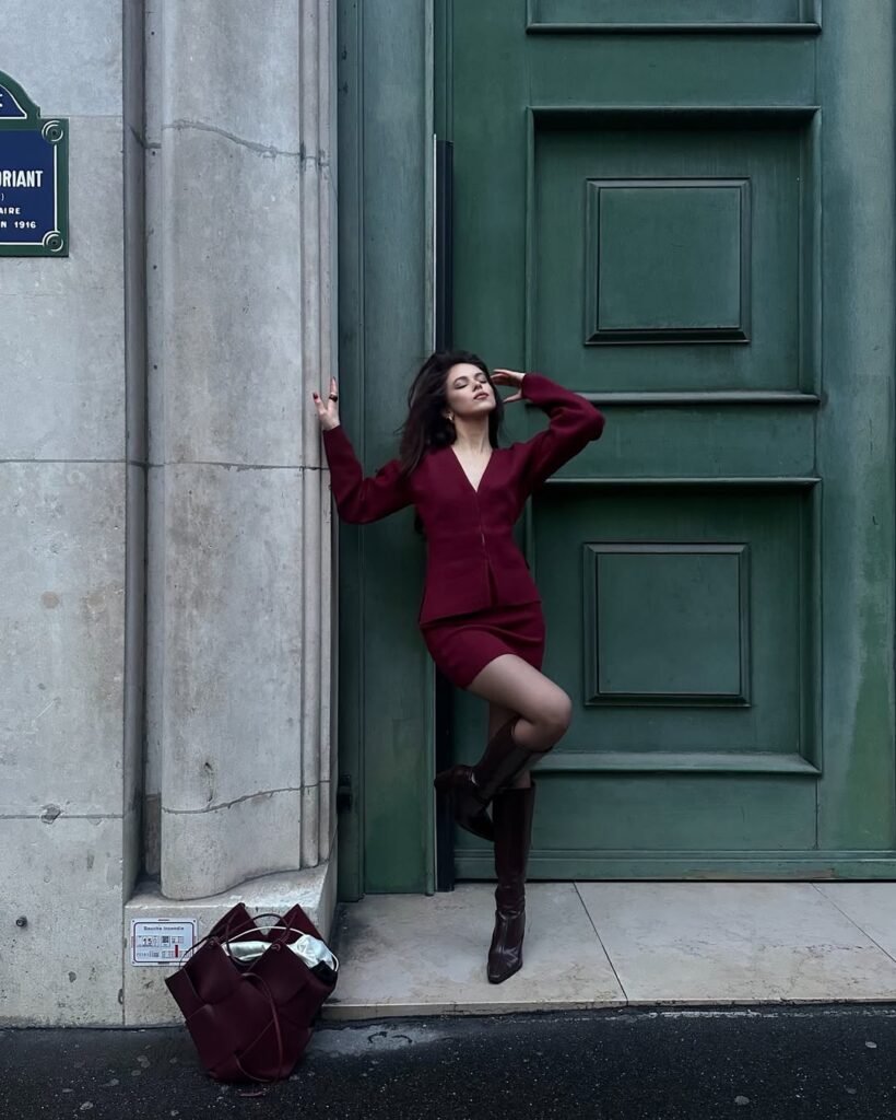 Woman in burgundy outfit with a fitted burgundy mini dress, matching jacket, and brown knee-high boots, posing against a large green door.