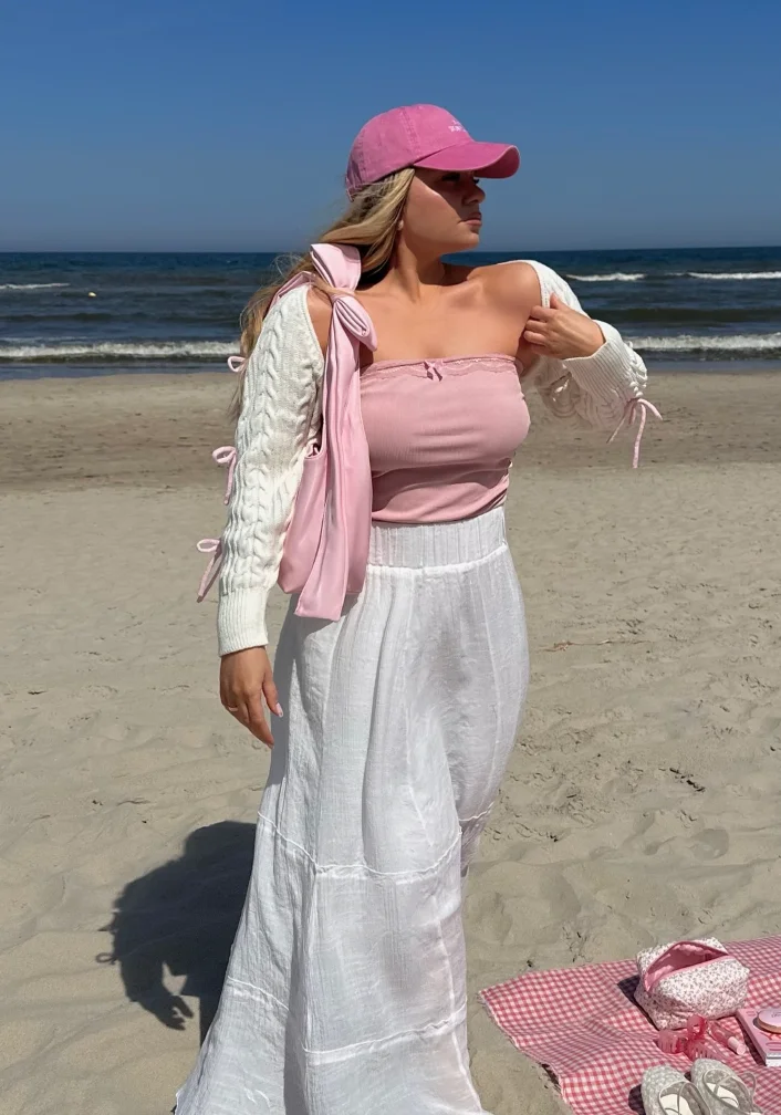 A woman on the beach in a strapless pink top, white wide-leg pants, a pink cap, and a knit cardigan, perfectly capturing cute simple outfits for coastal relaxation.