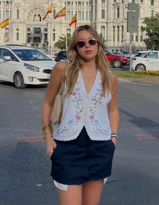 European Summer Outfit: White embroidered sleeveless blouse with black mini skirt, layered bracelets, and round sunglasses, posing in a city square.