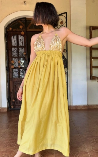 Woman in casual summer maxi dress with a mustard yellow skirt and sequined halter bodice, posing indoors near an open doorway.