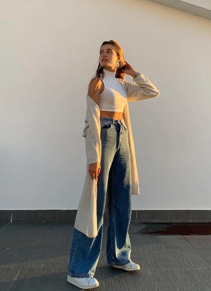 Woman in chill outfit ideas wearing a white sleeveless crop top, long beige cardigan, wide-leg blue jeans, and white sneakers, posing outdoors.
