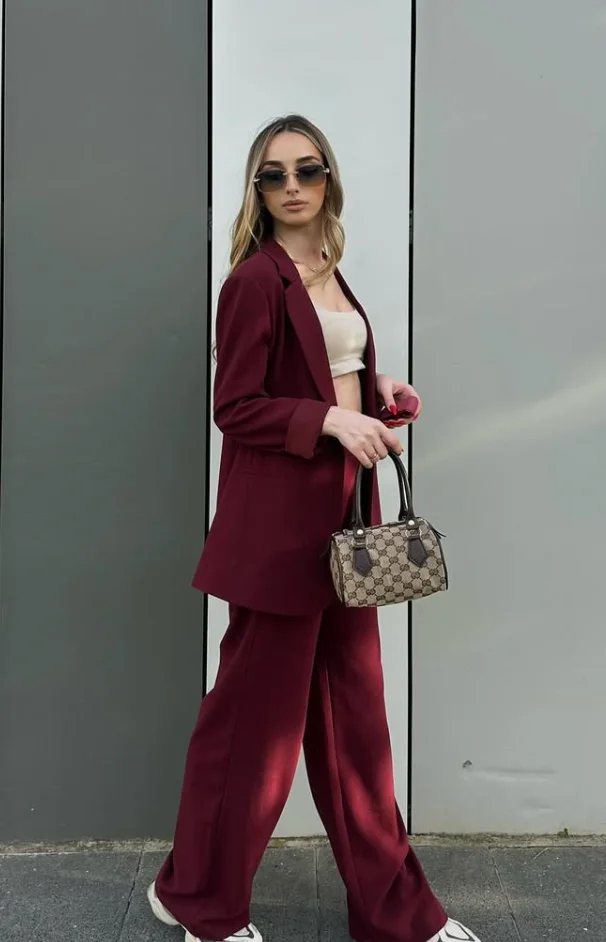 Woman in burgundy outfit wearing a burgundy oversized blazer, matching wide-leg pants, cream crop top, and white sneakers, holding a designer handbag.