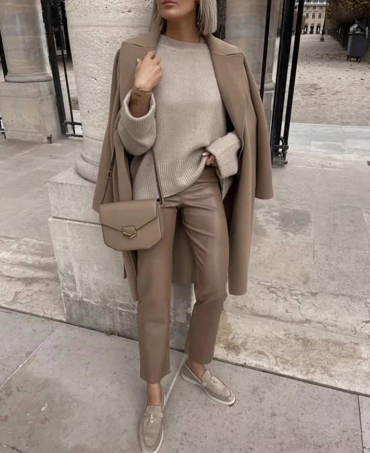 Brown Trousers Outfits – Woman dressed in beige leather cropped trousers, a beige knit sweater, and a matching overcoat draped over her shoulders, with a beige crossbody bag and loafers.