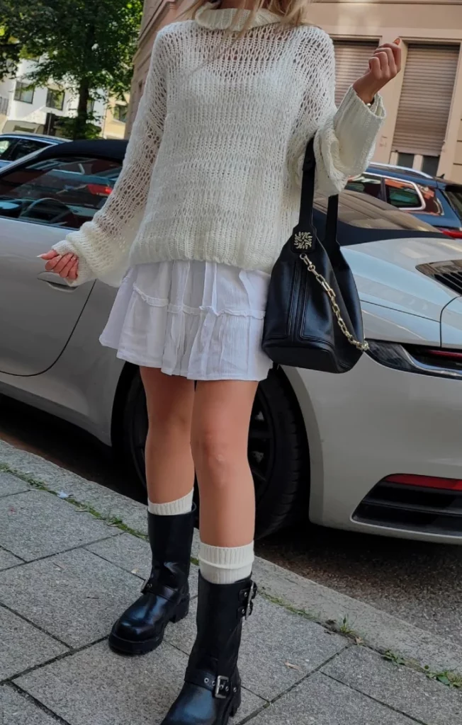 A woman wearing a see-through knit sweater over a short white skirt, paired with knee-high socks and black boots, offering an edgy take on cute simple outfits.