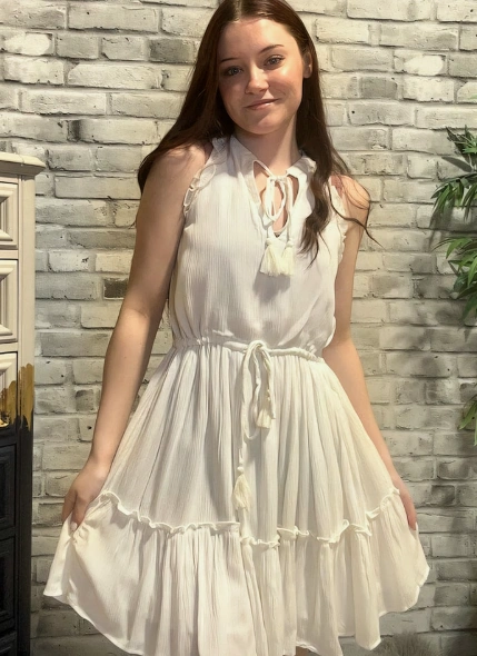 Woman wearing casual summer dress in plain white with a tie-front neckline and ruffled hem, posing indoors against a light brick wall.