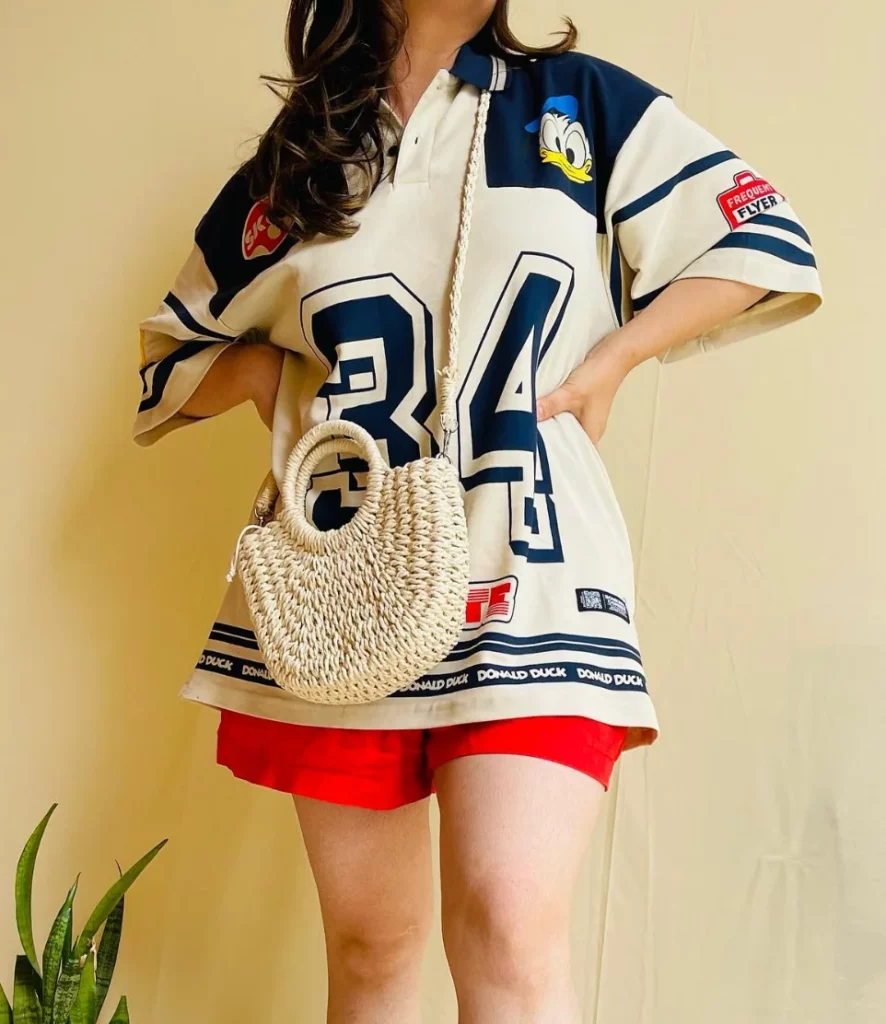 Woman in chill outfit ideas with an oversized Donald Duck sports jersey, bright red shorts, and a small woven handbag, standing indoors.