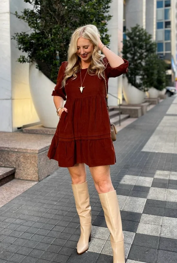 Woman in burgundy outfit featuring a short burgundy dress with ruffle details, beige knee-high boots, and a gold necklace, walking in the city.