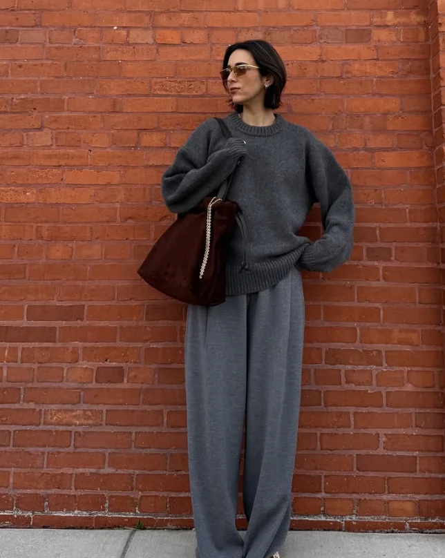 Relaxed grey trouser outfit with a loose dark grey sweater, oversized brown handbag, and white sneakers, styled against a brick wall.