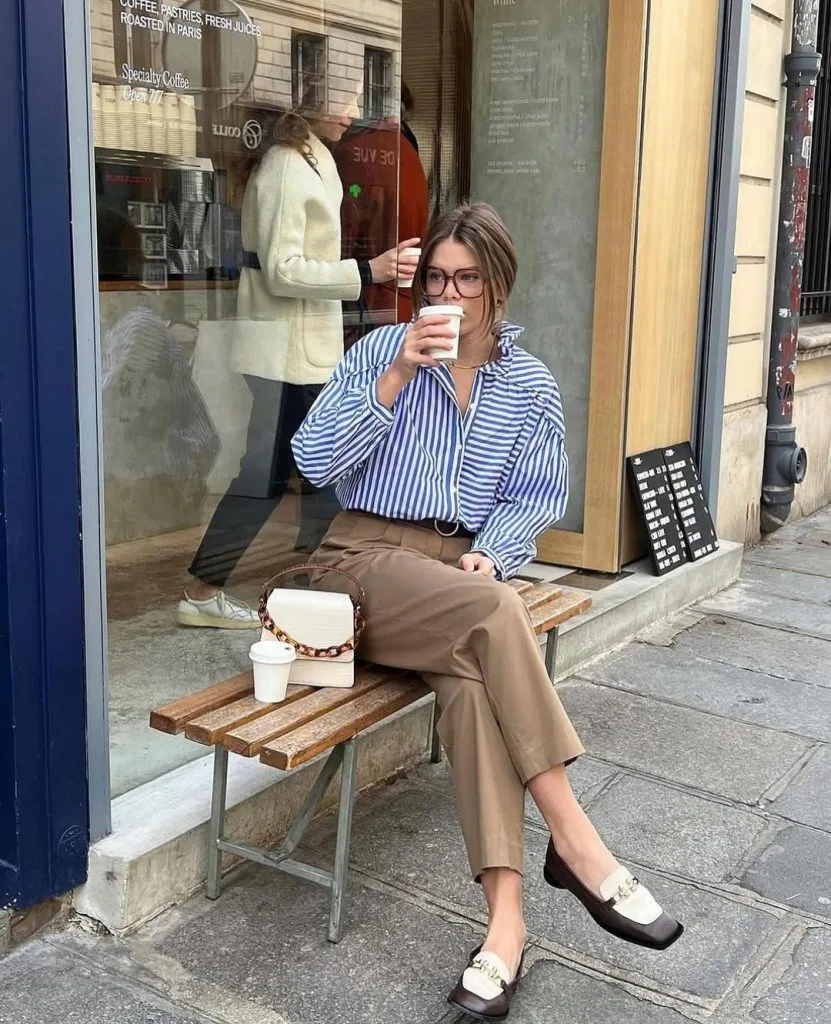 Office outfit with Parisian charm – blue-and-white striped shirt tucked into cropped camel trousers, paired with brown-and-white loafers and a chic cream handbag while enjoying a coffee break.