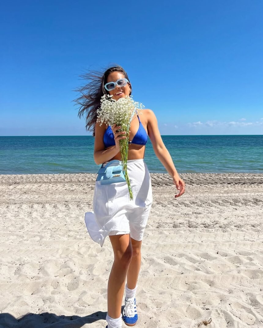 Woman posing on a sunny beach wearing a vibrant blue bikini top, white wrap skirt, and sneakers — a fun and stylish example of beach outfit ideas.