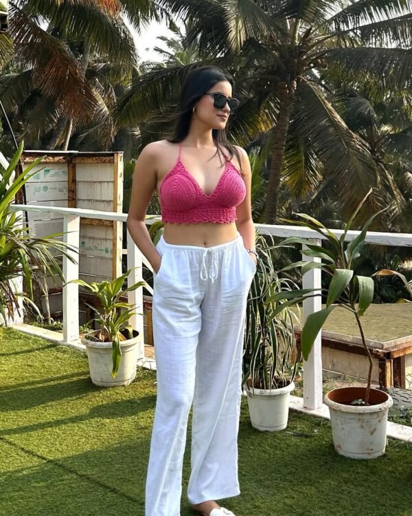A woman wearing a pink crochet halter top paired with white linen pants, sunglasses, and relaxed posture, enjoying a warm afternoon, ideal for a chic brunch in a tropical setting.