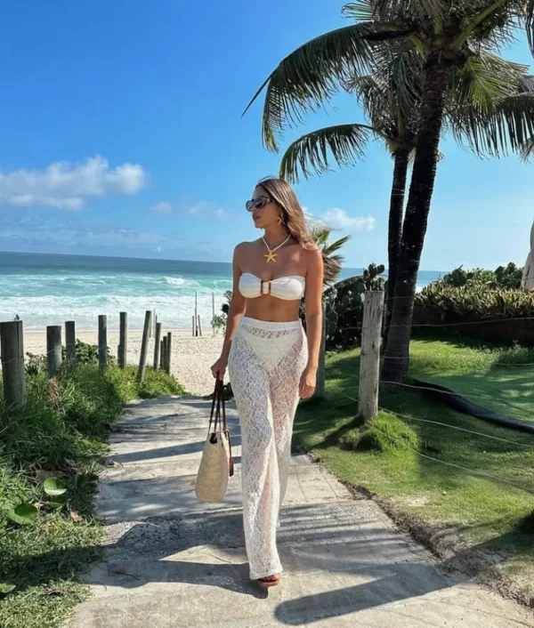Beach outfit with a white bandeau bikini top and sheer white lace pants, accessorized with a shell necklace, straw tote, and sunglasses on a sunny beachside path.