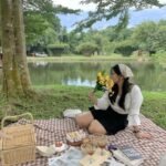 Picnic Date Outfits: Woman in a black skirt and white blouse with a beige headscarf holding flowers beside a lakeside picnic.