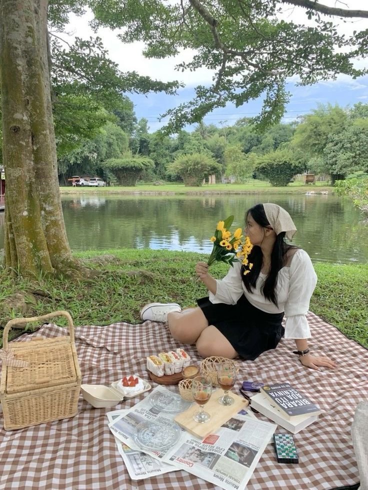 Picnic Date Outfits: Woman in a black skirt and white blouse with a beige headscarf holding flowers beside a lakeside picnic.