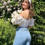 Woman wearing a floral and denim blazer outfit with a white off-shoulder top, standing near blooming bushes.