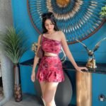 Woman wearing a red patterned one-shoulder crop top paired with a matching skirt, standing indoors by a decorative table, showcasing trendy skirt outfits.