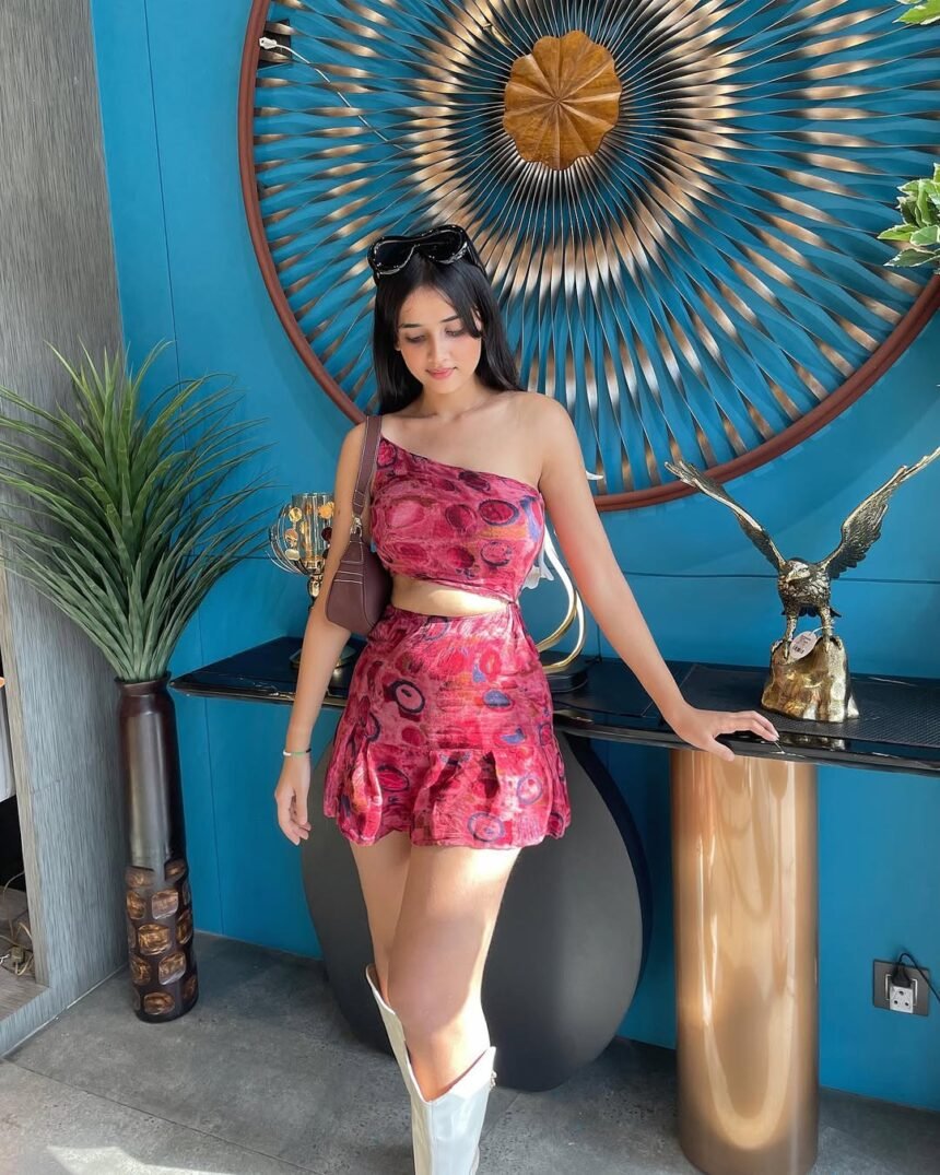 Woman wearing a red patterned one-shoulder crop top paired with a matching skirt, standing indoors by a decorative table, showcasing trendy skirt outfits.
