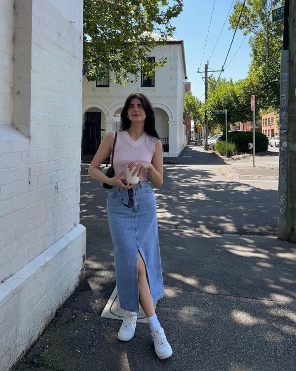 A woman standing on a quiet street corner wearing a light pink sleeveless top and a front-slit blue denim midi skirt, paired with white sneakers — Casual and comfy Skirt Outfits for everyday errands.