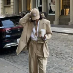 Woman in a camel oversized blazer over white shirt and tan wide-leg pants, accessorized with a tan cap and coffee cup, walking on a city street. Blazer Outfits.