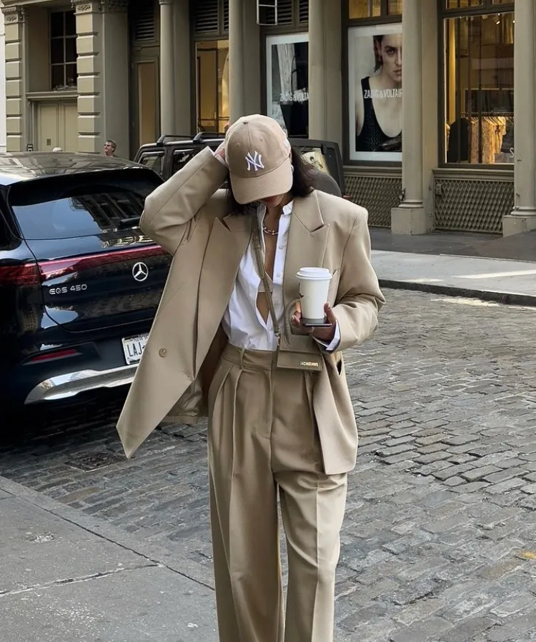 Woman in a camel oversized blazer over white shirt and tan wide-leg pants, accessorized with a tan cap and coffee cup, walking on a city street. Blazer Outfits.