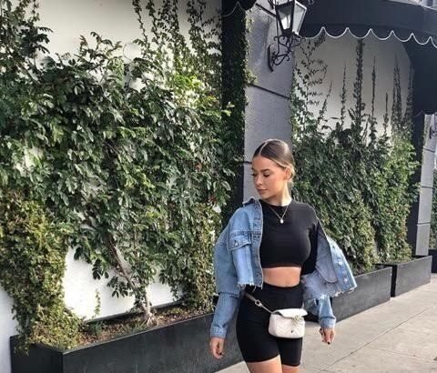 Woman walking on the street wearing black biker shorts outfit paired with a black crop top, light blue denim jacket, white sneakers, and a white waist bag. Stylish Biker Shorts outfits.