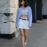 A woman in a structured mini white skirt with a black belt, cropped knit top, and a striped blue button-down, paired with Mary Jane flats – a modern preppy old money spring outfit.