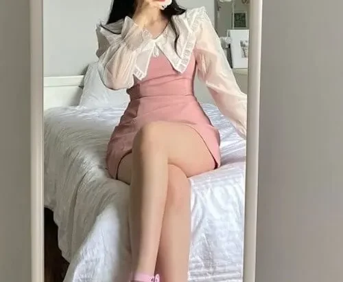 Coquette outfit ideas in a pastel pink pinafore-style mini dress layered over a white sheer puff-sleeve blouse with a pointed collar, paired with matching pink heels and a headband.