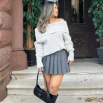 Casual Thanksgiving outfit featuring a gray off-shoulder knit sweater paired with a pleated gray mini skirt, knee-high black boots, and a black handbag.