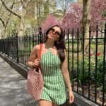 Woman in casual summer dress featuring green and white vertical stripes, halter neckline, paired with sunglasses, a pink woven tote bag, and walking in a sunny park with blooming trees.