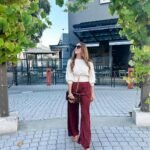 Woman in burgundy outfit featuring wide-leg burgundy pants, a white sweater, tan heels, and a brown handbag, walking outdoors.