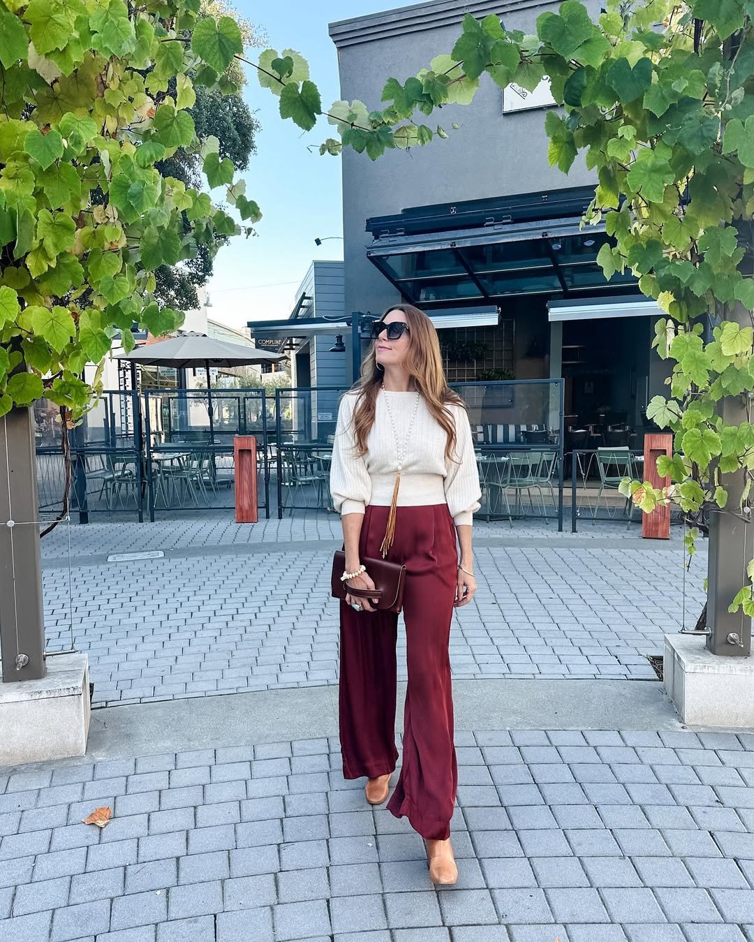 Woman in burgundy outfit featuring wide-leg burgundy pants, a white sweater, tan heels, and a brown handbag, walking outdoors.