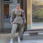 Woman in chill outfit ideas with a matching taupe tracksuit, tie-dye crop top, and white sneakers, standing on a city sidewalk.