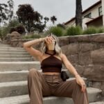 Brown Trousers Outfits – Woman sitting on outdoor stairs wearing light brown wide-leg trousers, a dark brown halter knit crop top, and white sneakers, holding an iced coffee in front of her face.