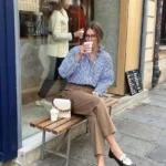 Office outfit with Parisian charm – blue-and-white striped shirt tucked into cropped camel trousers, paired with brown-and-white loafers and a chic cream handbag while enjoying a coffee break.